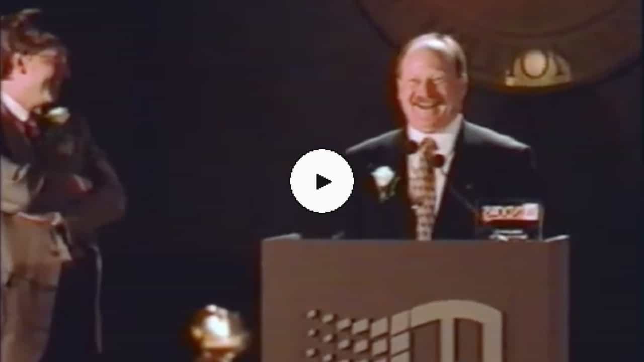 A picture of Alan Cooper in a suit smiling and speaking while he receives an award. Bill Gates is looking on from the side, also smiling. There's a play button overlayed on top.